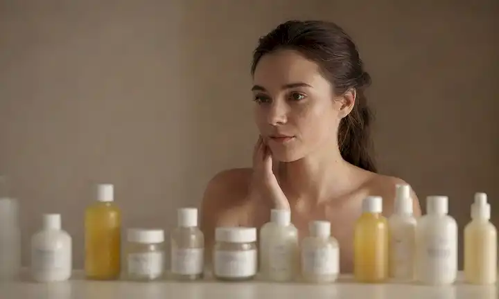Una persona tranquila rodeada de botellas y jarras de productos de cuidado de la piel, con un brillo sutil de luz; enfoque suave en la textura de la piel y los poros; sombras suaves para transmitir serenidad y relajación; reflejos mínimos en superficies; colores naturales y texturas; sin iluminación dura o composiciones ocupadas