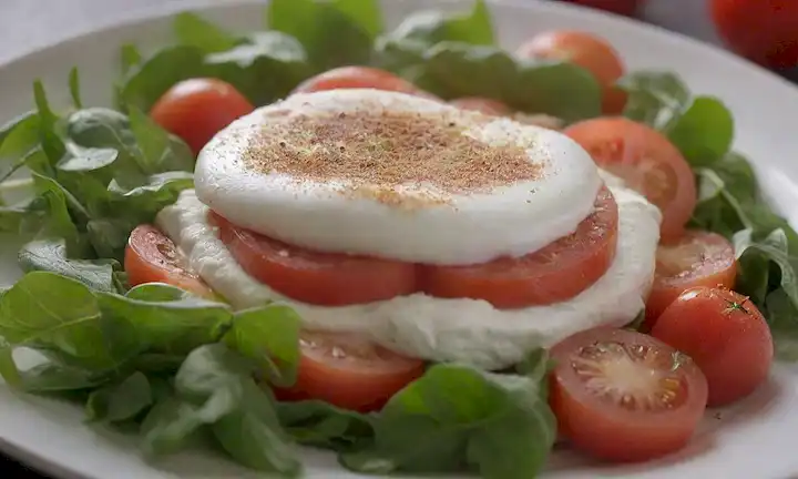 Una placa de tono cálido con una corteza dorada, rematada con un delicado puré de tonos blancos y carmesí cremosos, rodeada de una dispersión de verdes frescos y tomates jugosos rebanados, adornados con unos pocos wisps de tiernas cadenas de mozzarella búfalo