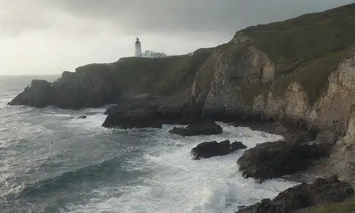 Una foto de una costa escarpada y barrida, con afloramientos rocosos, ondas de choque, y posiblemente un faro u otra estructura costera, con un indicio de maldad en el aire, transmitiendo una sensación de salvaje y aislamiento